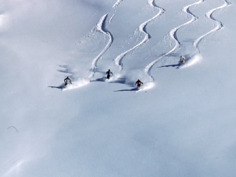 Quatre skieurs dévalant une pente enneigée, laissant des traces parallèles dans la neige fraîche.