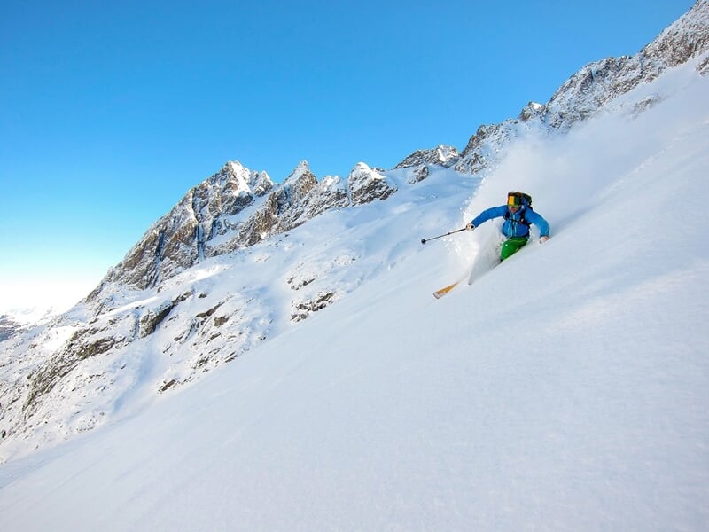 Skieur en tenue bleue et verte descendant une pente enneigée avec des montagnes en arrière-plan.