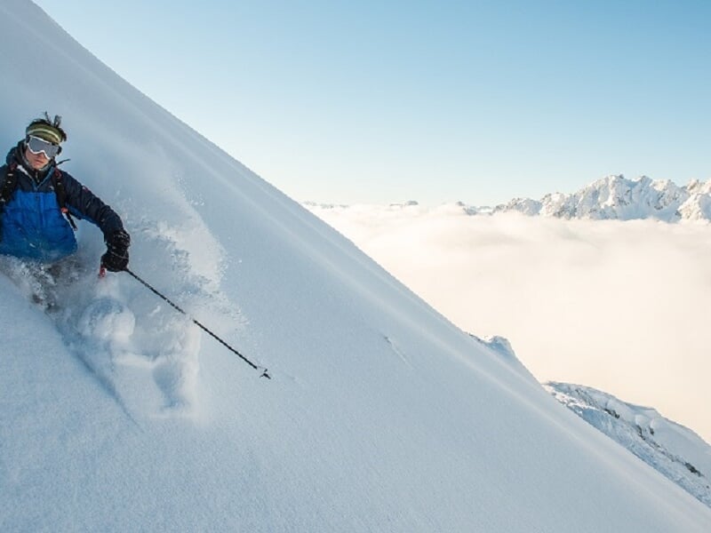 Skieur en bleu dévalant une pente enneigée avec un ciel bleu et un paysage de montagnes en arrière-plan.