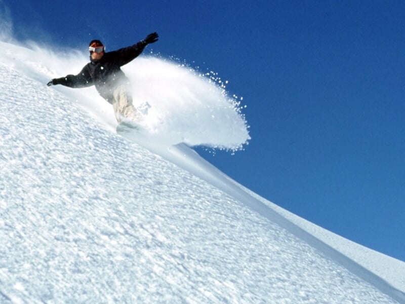 Snowboardeur effectuant une descente rapide sur une pente enneigée sous un ciel bleu clair.