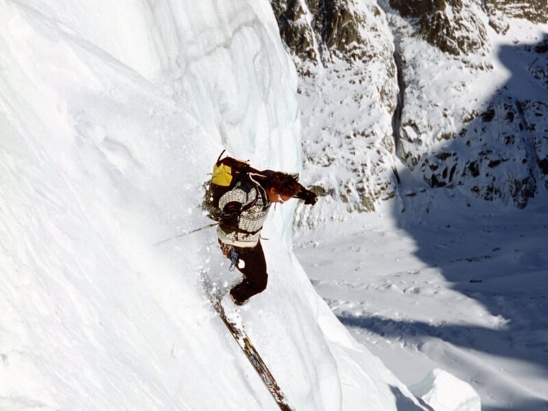 Skieur en tenue claire descendant une pente raide et enneigée, entouré de rochers et de neige.