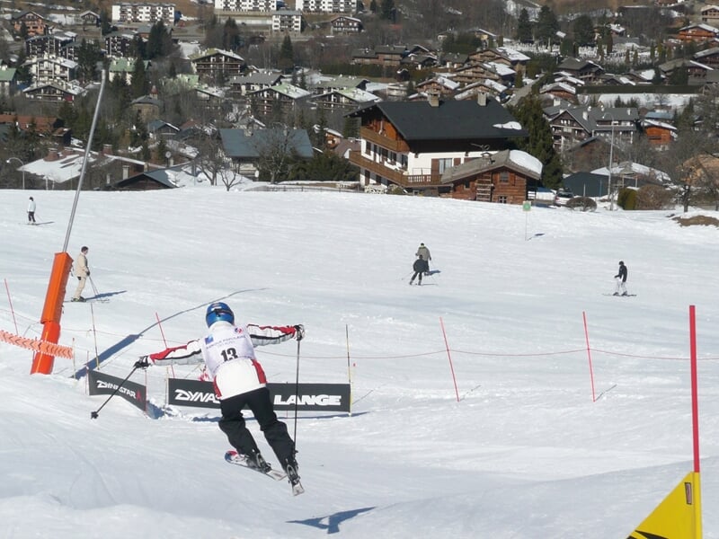 Skieur descendant une piste de ski avec des barrières et un village en arrière-plan sous un ciel clair.