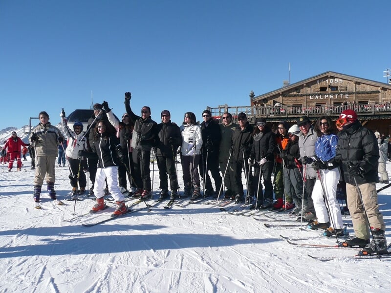 Groupe de skieurs posant ensemble sur une piste enneigée devant un chalet en montagne par beau temps.