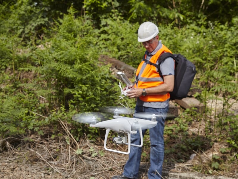 Homme en gilet orange et casque blanc préparant un drone blanc au sol dans un environnement boisé.