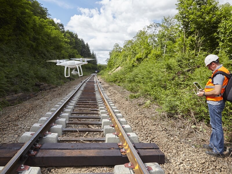 Homme contrôlant un drone volant au-dessus d'une voie ferrée entourée d'arbres verts.
