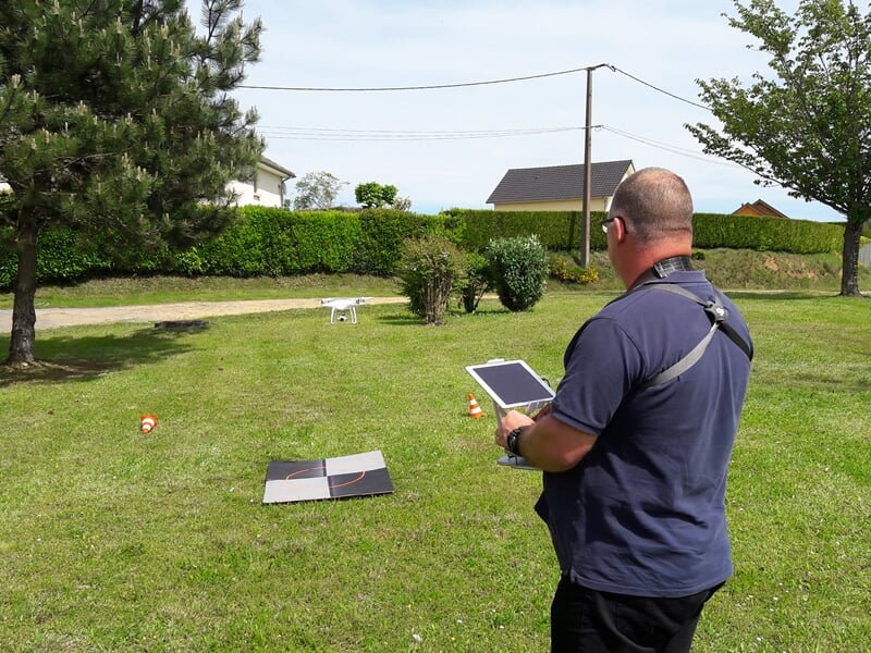 Personne pilotant un drone blanc dans un jardin avec une tablette en main et des cibles au sol.