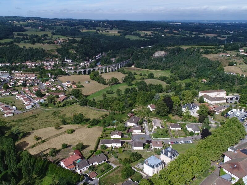 Vue aérienne d'un village avec des maisons, des arbres et un viaduc dans un paysage verdoyant.