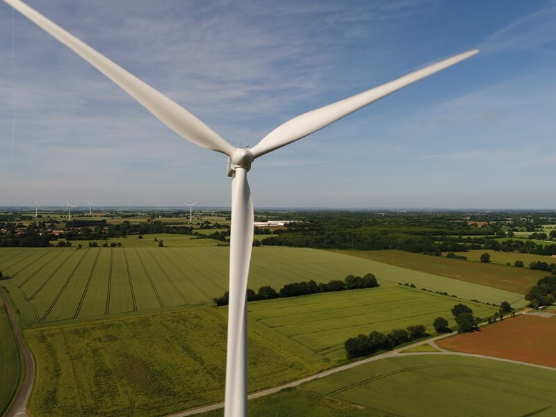 Gros plan aérien d'une éolienne blanche au-dessus de champs cultivés et d'une forêt lointaine.