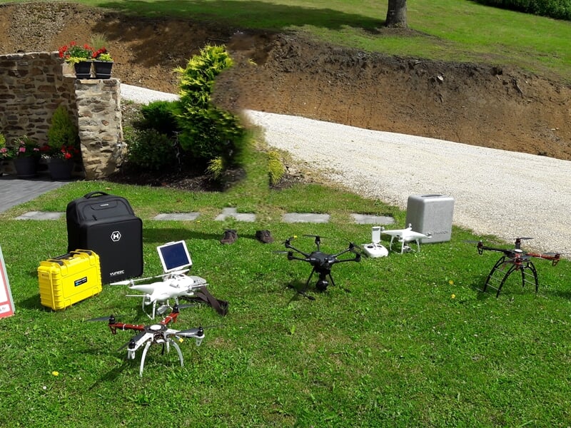 Plusieurs drones et équipements de pilotage posés sur une pelouse verte près d'un mur en pierre.
