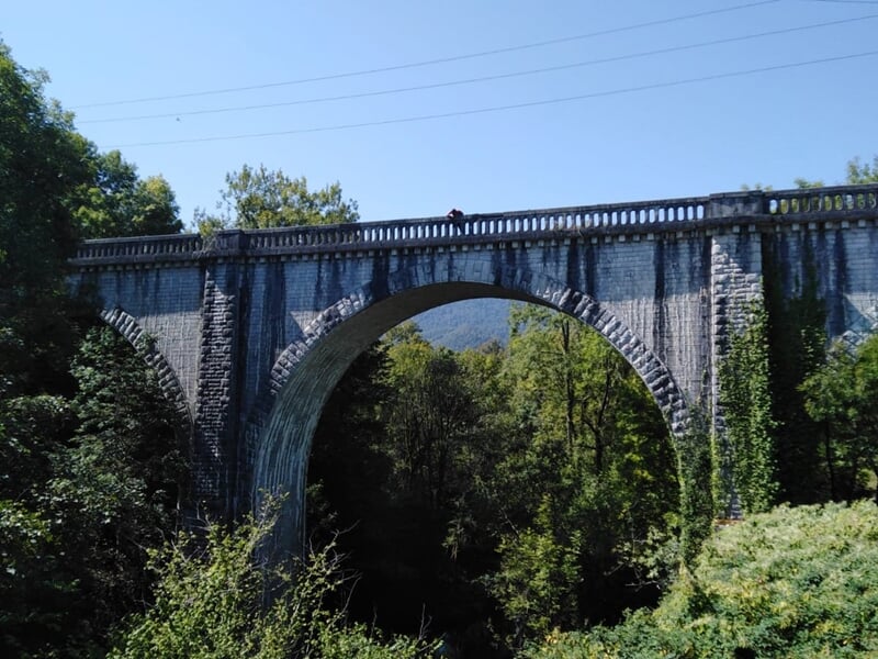 Un pont en pierre avec une arche large, entouré de végétation dense sous un ciel bleu clair.