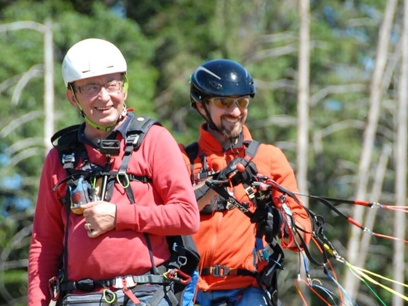 Deux hommes souriants équipés pour un baptême de parapente, l'un portant un casque blanc et l'autre un casque noir.