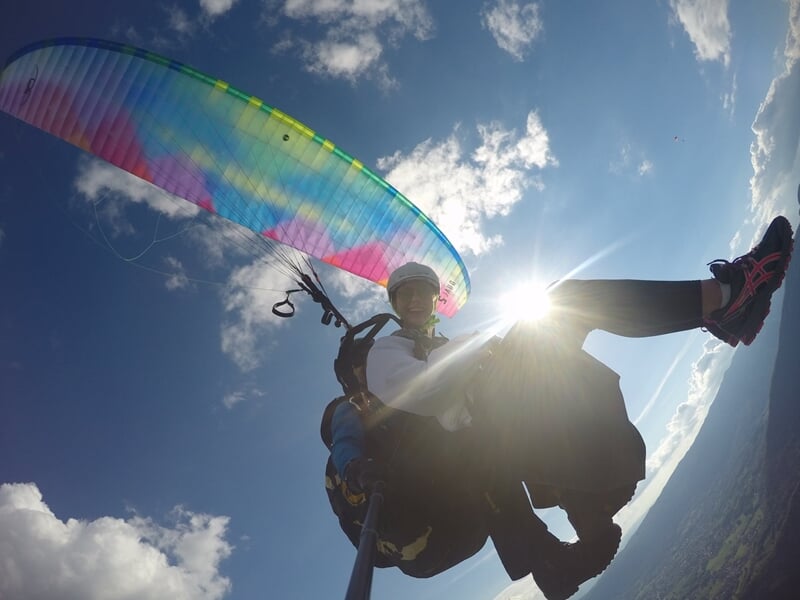 Personne en vol en parapente avec une voile colorée, le soleil brillant derrière et un ciel partiellement nuageux.
