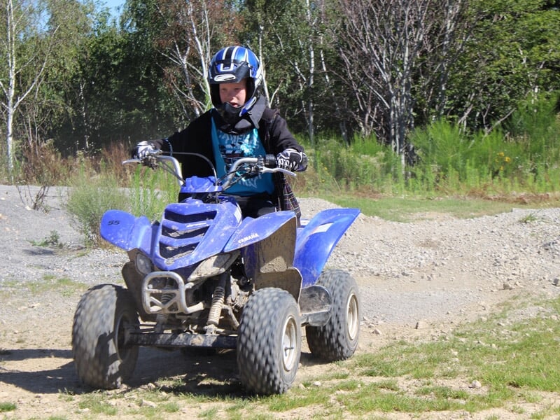 Jeune garçon en casque bleu conduisant un quad bleu sur un chemin de terre entouré d'arbres verts en été