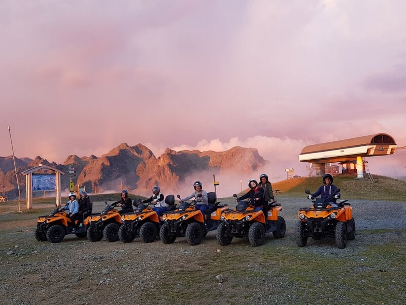 Groupe de personnes en randonnée sur des quads orange au sommet d'une montagne avec un paysage de montagnes au coucher du soleil.