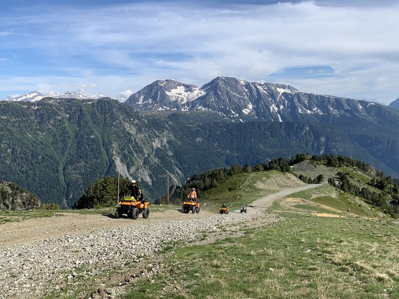 Quatre personnes en randonnée sur des quads sur un chemin de montagne à Chamrousse, avec un paysage alpin enneigé en arrière-plan.