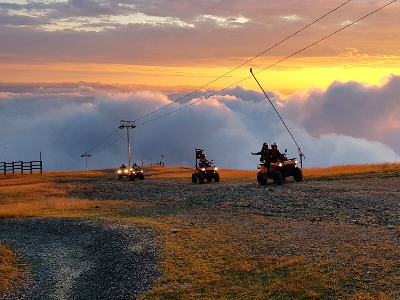 Quatre personnes en randonnée en quad sur un chemin de montagne au coucher du soleil avec un ciel nuageux spectaculaire.