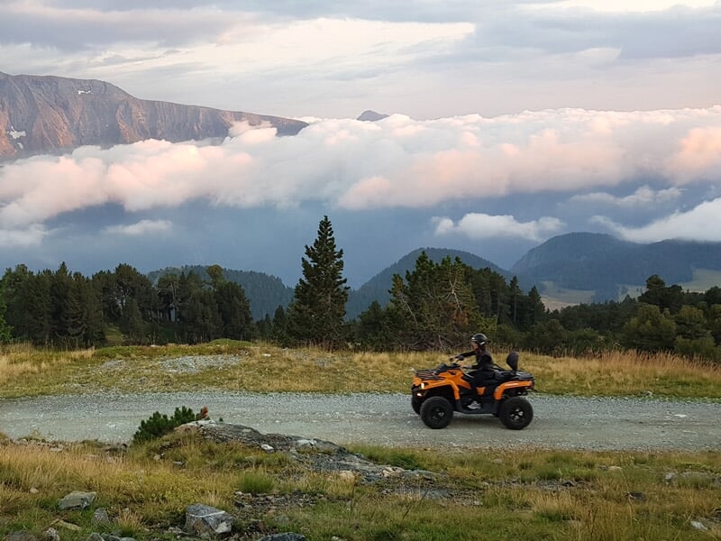 Personne conduisant un quad orange sur un chemin de montagne avec forêt et nuages en arrière-plan au coucher du soleil.