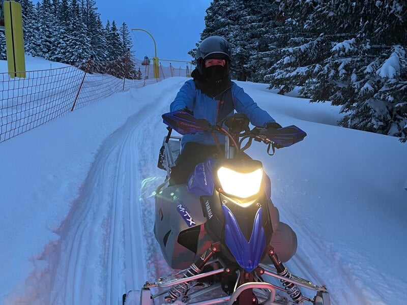 Personne en tenue d'hiver conduisant une motoneige bleue sur un sentier enneigé en forêt.