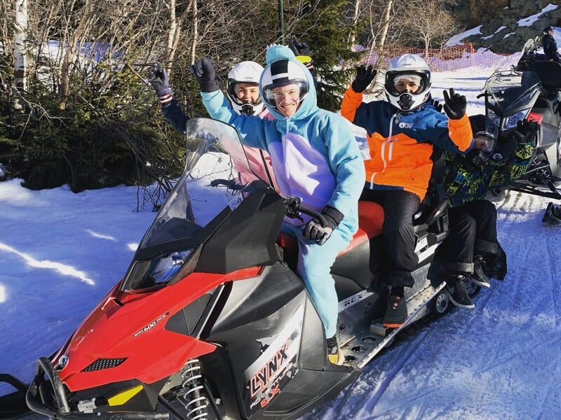 Trois personnes souriantes sur une motoneige rouge dans un paysage enneigé et ensoleillé.