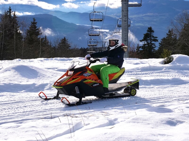Personne en tenue d'hiver conduisant une motoneige orange sur un terrain enneigé en montagne.