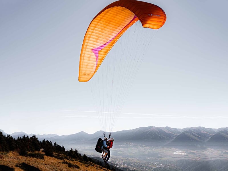 Personne en parapente avec voile orange survolant un paysage montagneux au coucher du soleil, ambiance paisible et claire.