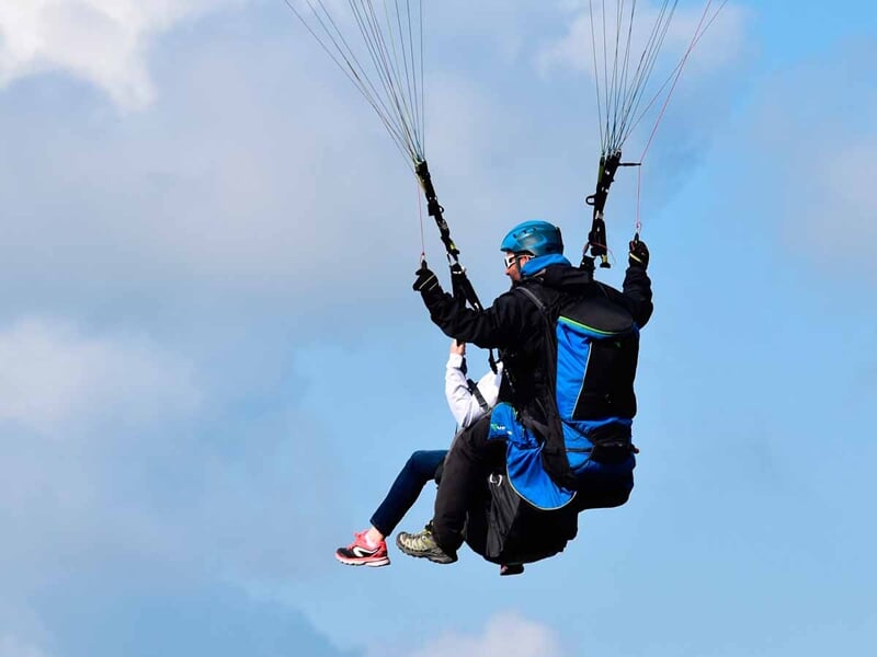 Vol en tandem de parapente avec deux personnes équipées de casques, dans un ciel bleu clair avec quelques nuages.