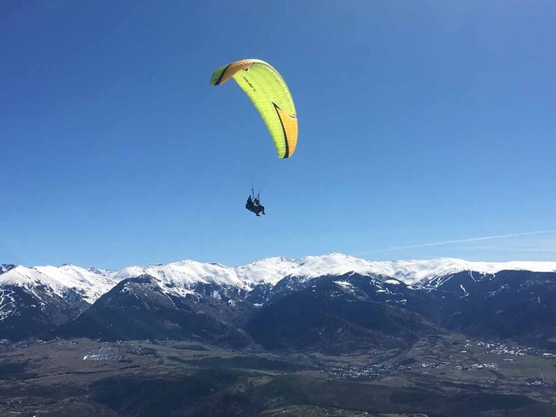 Parapente jaune avec deux pilotes volant au-dessus d'une vallée entourée de montagnes enneigées sous un ciel bleu.