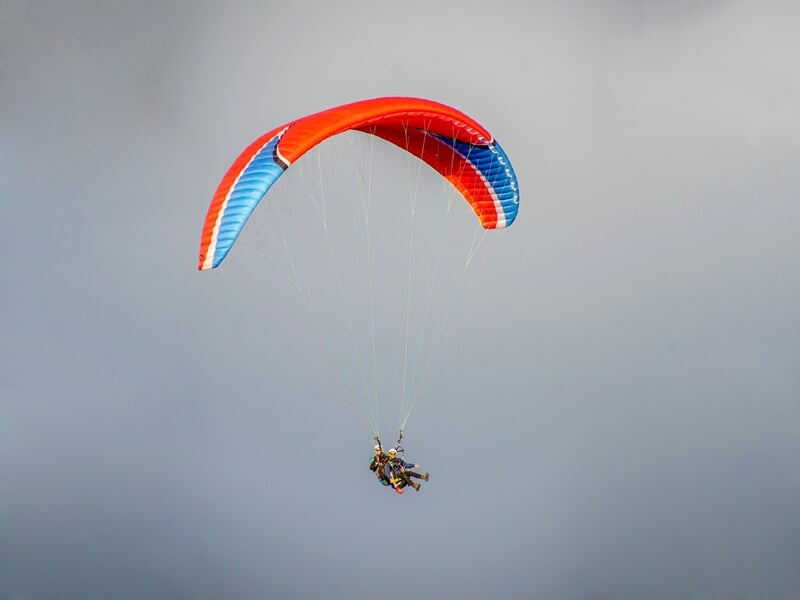 Deux personnes en parapente avec voile rouge et bleue, volant dans un ciel nuageux avec un fond de montagnes.