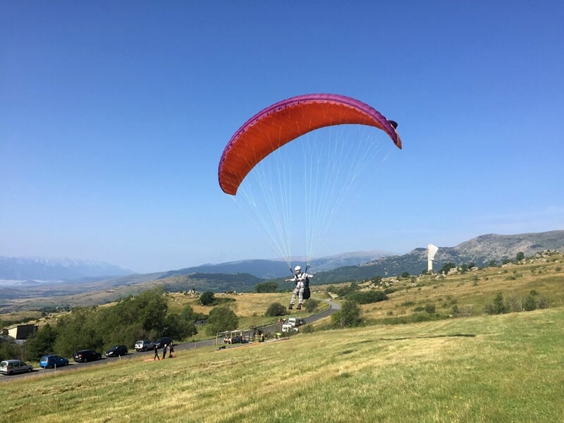 Parapente rouge avec pilote en train d'atterrir sur une prairie verte avec des collines et des voitures en arrière-plan.