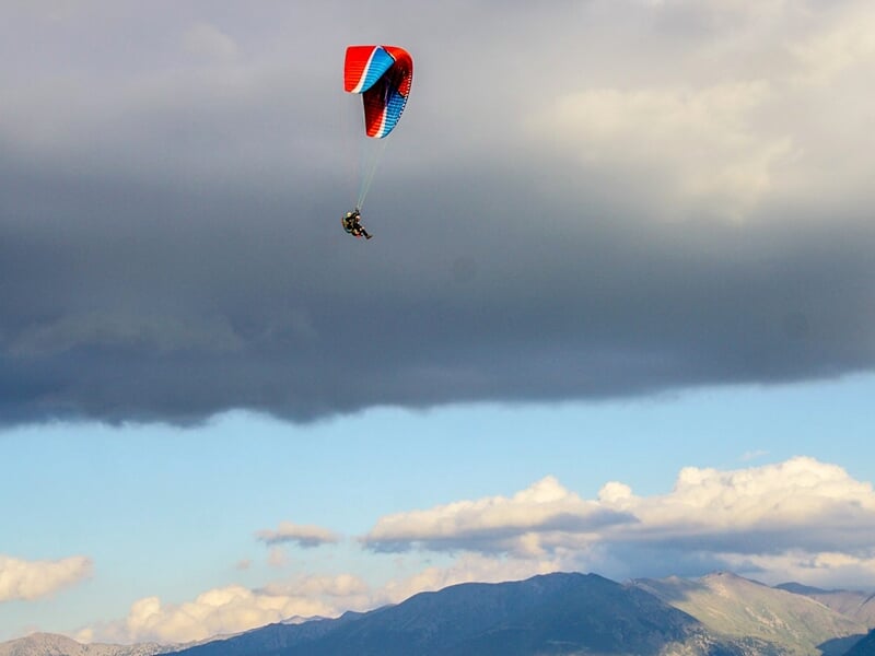 Parapente rouge et bleu avec deux personnes volant haut dans un ciel nuageux au-dessus d'un paysage montagneux.