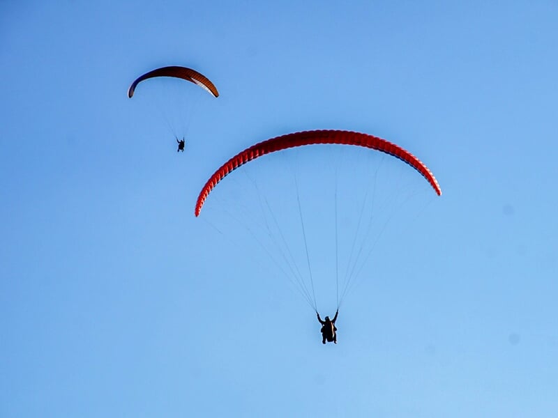 Deux parapentes rouges volant dans un ciel bleu clair, silhouettes visibles avec montagnes en arrière-plan.