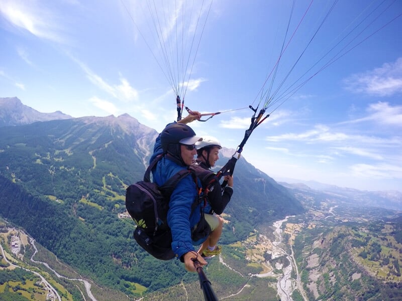 Deux personnes en parapente survolant une vallée verdoyante et des montagnes majestueuses sous un ciel bleu clair avec des nuages épars.