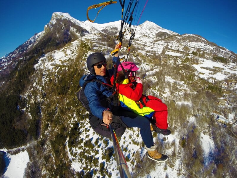 Parapentiste et enfant en vol tandem au-dessus de montagnes enneigées avec un ciel bleu clair, l'enfant portant une combinaison rouge et un casque rose.