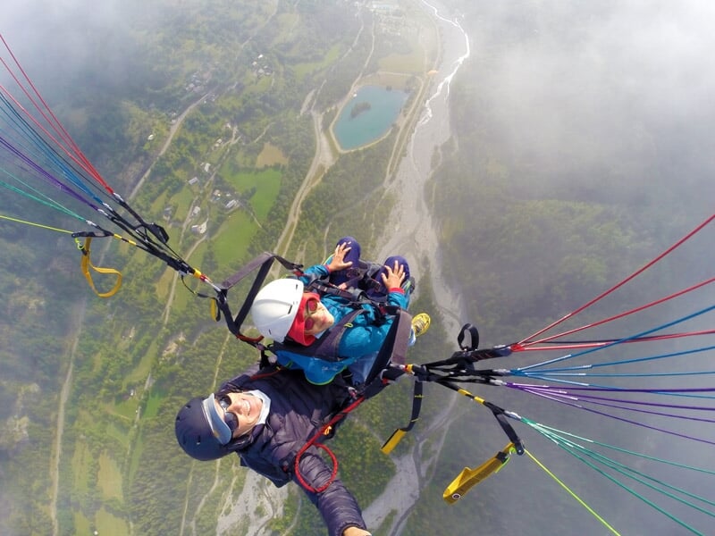 Parapentiste en tandem survolant un paysage de montagnes et de rivières, partiellement masqué par des nuages, avec des câbles multicolores du parapente visibles.