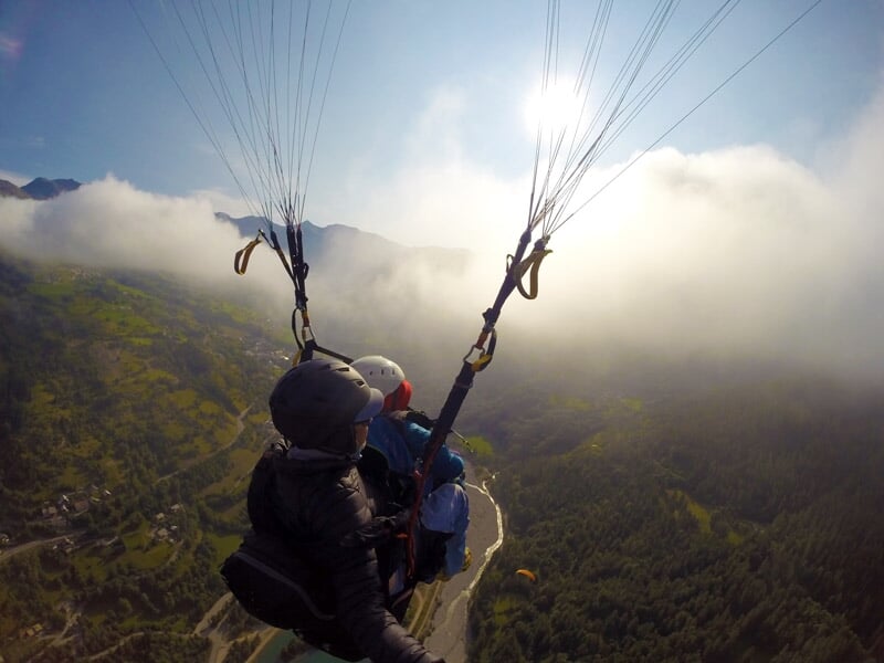 Vue arrière d'un vol en parapente au-dessus d'une vallée montagneuse verdoyante avec le soleil éclatant à travers les nuages à l'horizon.