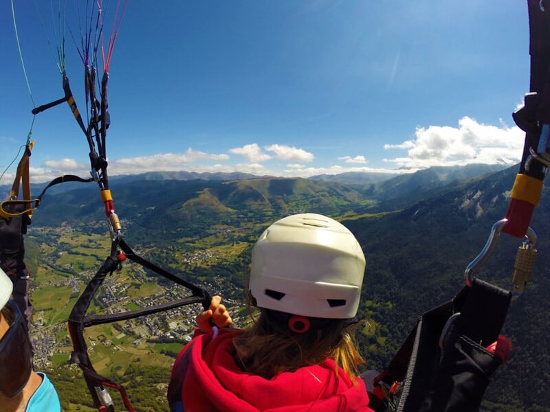 Vue arrière d'un passager en parapente regardant un paysage de vallée et montagnes verdoyantes sous un ciel bleu avec des nuages blancs dispersés.