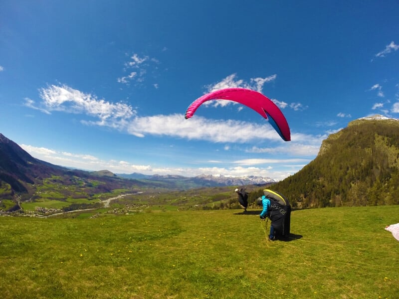Parapentistes en phase de décollage sur une pente herbeuse avec un parapente rose déployé sous un ciel bleu clair et des montagnes en arrière-plan.