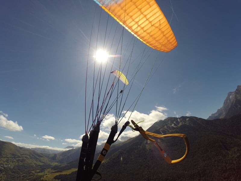 Parapente orange illuminé par le soleil, en vol au-dessus d'une vallée verdoyante, montagnes en arrière-plan, sous un ciel bleu