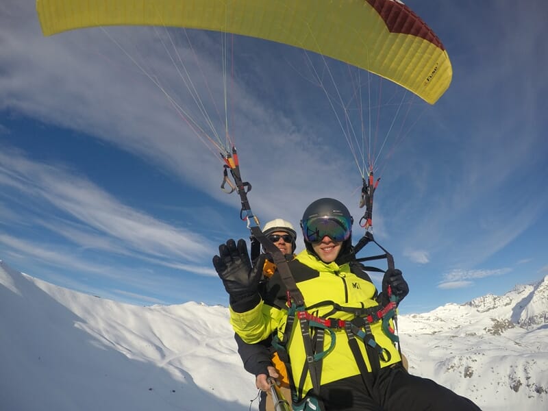 Parapentiste en combinaison jaune volant au-dessus d'une piste de ski, montagnes en arrière-plan, ciel bleu