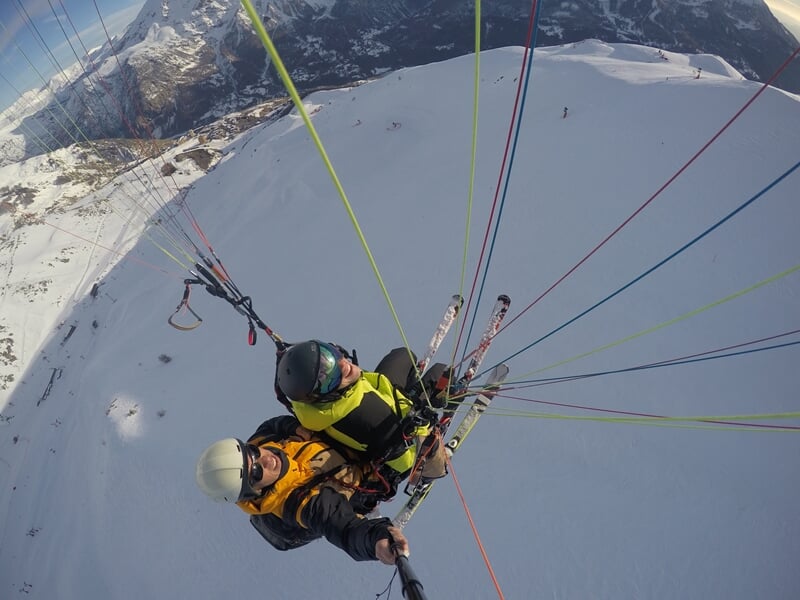 Duo de parapentistes en vol au-dessus d'une piste de ski, chaîne de montagnes à l'horizon, ciel bleu