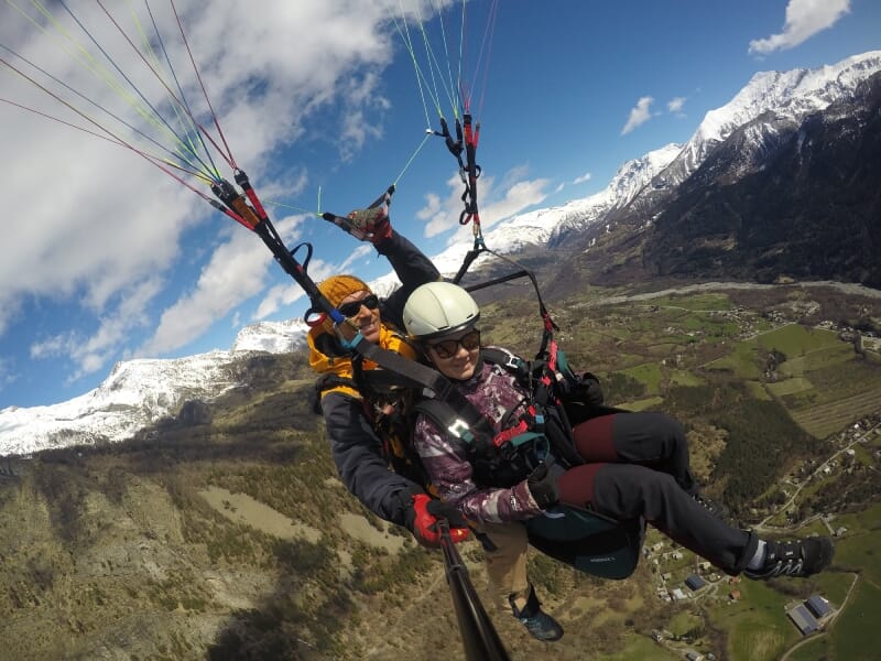 Vol en parapente en tandem au-dessus de la vallée, pilote en combinaison orange et passager en veste violette, montagnes enneigées et villages en arrière-plan.