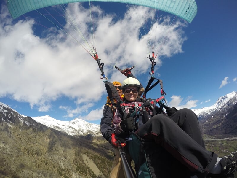 Vol en parapente en tandem, pilote en combinaison orange et passager en veste violette, ciel bleu avec quelques nuages, montagnes enneigées en arrière-plan.
