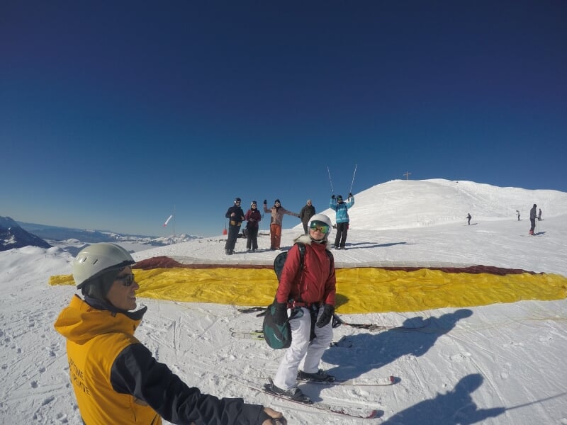 Groupe de skieurs au sommet d'une montagne enneigée, préparant un vol en parapente, voile jaune déployée au sol, ciel bleu dégagé.