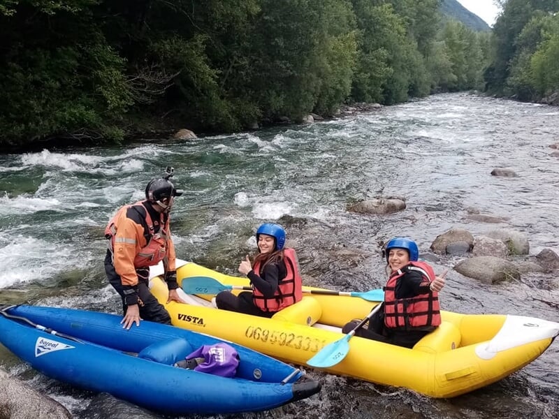 Trois personnes en gilets de sauvetage et casques bleus prêtes à faire du rafting sur une rivière en montagne.