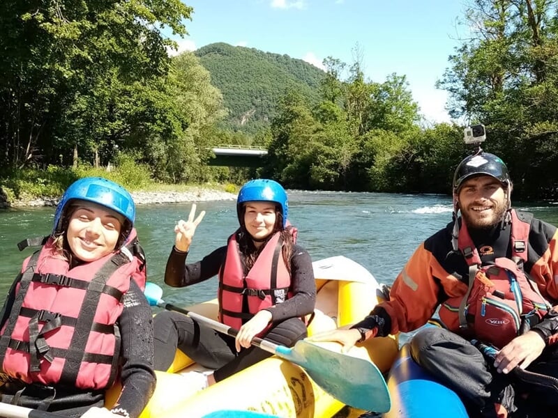 Trois personnes souriantes en gilets de sauvetage et casques bleus sur un raft jaune sur une rivière calme.