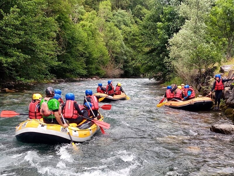 Groupe de six personnes en raft jaune avec casques bleus et gilets rouges pagayant sur une rivière en forêt.