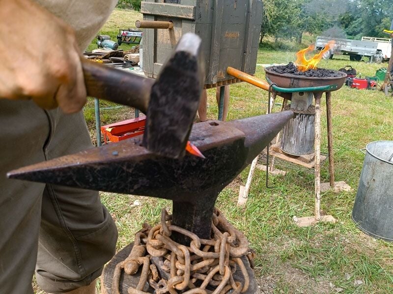 Personne forgeant un outil en métal rougeoyant sur une enclume entourée d'une chaîne en extérieur vert.