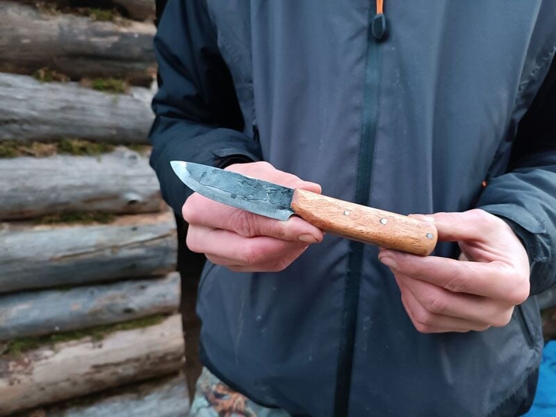 Personne tenant un couteau artisanal avec un manche en bois devant un mur en rondins de bois.