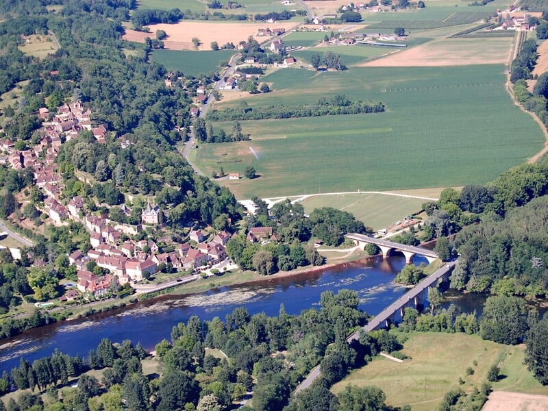 Petit village au bord d'une rivière avec un pont ancien et des champs verts environnants.