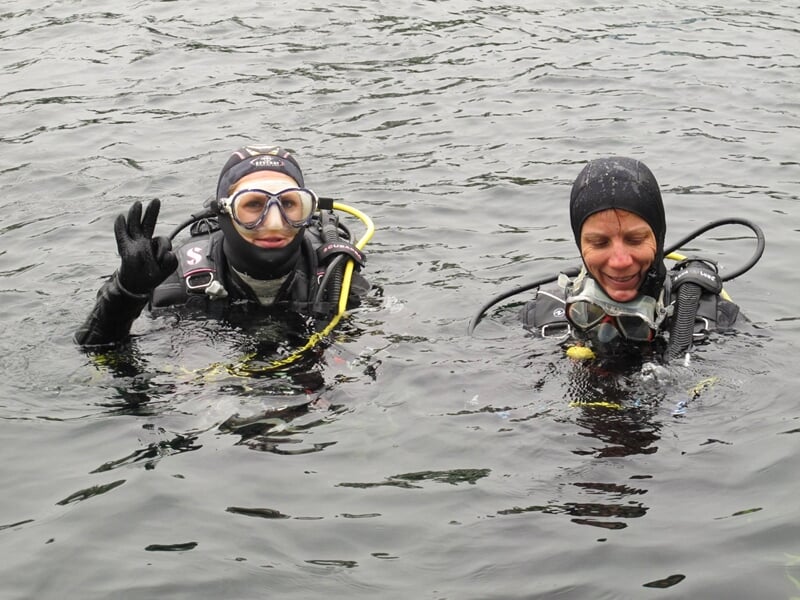 Deux plongeurs en combinaison et masque font un signe de la main dans une eau calme lors d'un baptême de plongée.